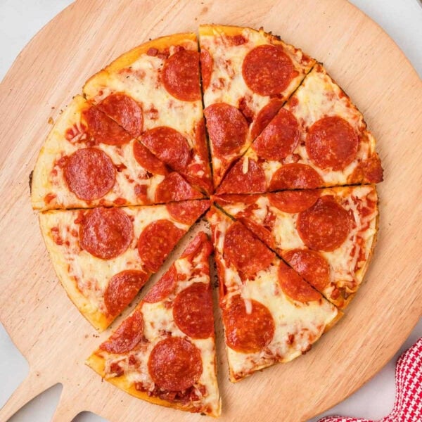 A whole pepperoni pizza, cooked like a Frozen Pizza on Blackstone and cut into eight slices, is displayed on a wooden pizza board. The golden crust, melted cheese, and pepperoni slices shine next to a red-and-white cloth in the corner.