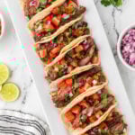 Five Blackstone Steak Tacos topped with diced tomatoes, red onions, and chopped cilantro are arranged on a rectangular white platter. Lime slices, a bowl of onions, and a striped napkin are nearby on a marble surface.