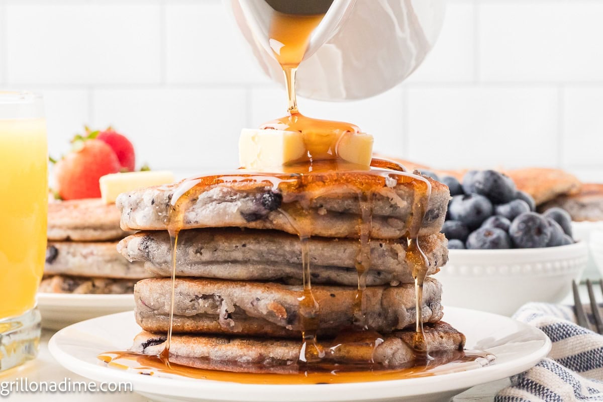 Blueberry Pancakes stacked on a white plate topped with maple syrup and butter