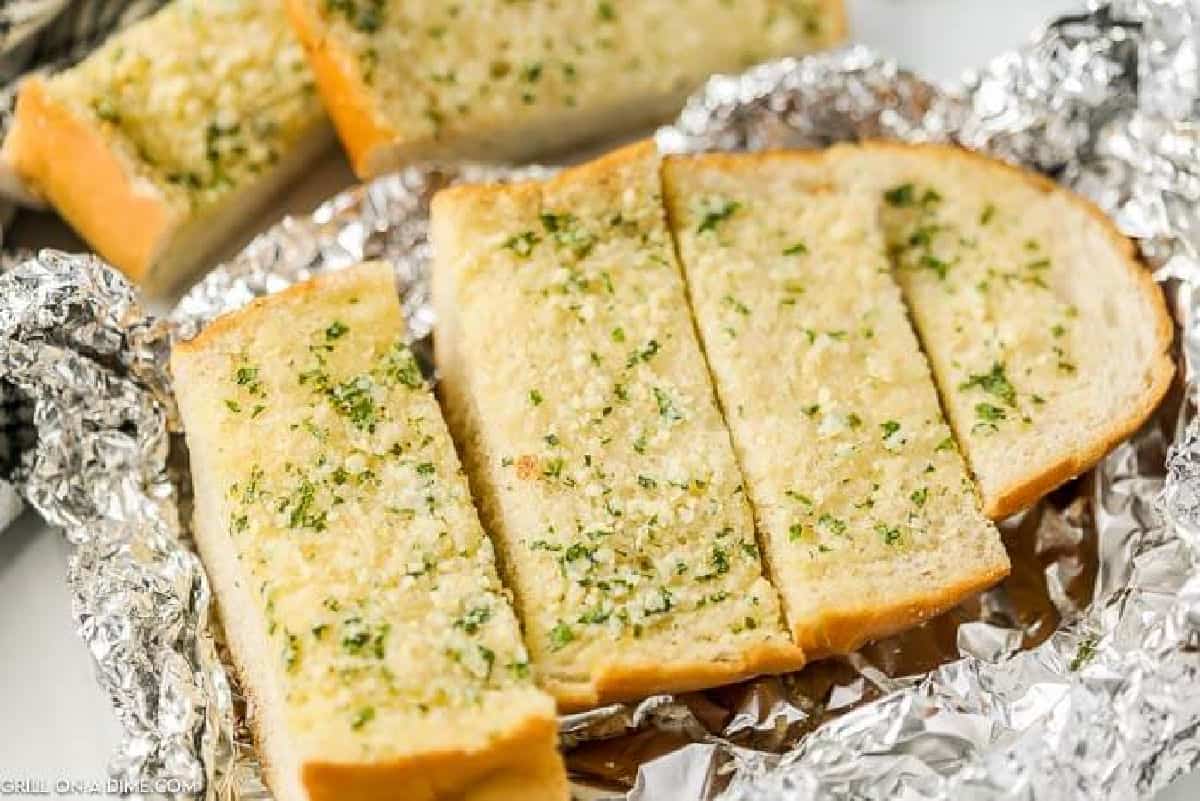 Garlic bread slices with herbs on top, resting on crinkled aluminum foil, ready to serve.