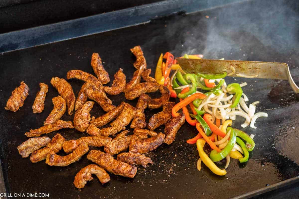 Cooked Steak Fajitas on a griddle on green, red, yellow peppers strips with onions cooking on the griddle.