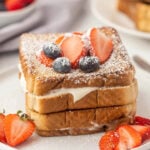 A stack of stuffed French toast slices filled with cream, topped with powdered sugar, sliced strawberries, and blueberries, served on a white plate with extra strawberries on the side.