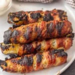 Grilled bacon-wrapped pickles on a white plate, fresh and smoky for appetizers.