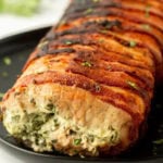 Seasoned bacon-wrapped stuffed pork loin on black plate, cooked to perfection with herbs.