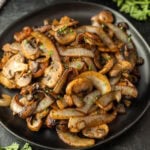 A black plate filled with Blackstone Sautéed Mushrooms and Onions for Steak, garnished with chopped herbs, sits on a dark surface. The caramelized vegetables glisten, with fresh parsley visible in the background.