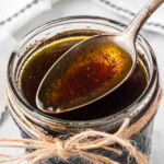 Close-up of a glass jar filled with a dark liquid, possibly a Hibachi Steak Marinade or balsamic vinaigrette, with a spoon resting on top. The jar is decorated with a rustic twine bow around its neck.