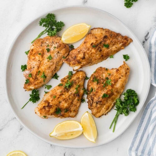 Blackstone Catfish on a white plate with fresh herbs and lemon wedges on the side