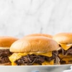 Three Onion Smash Burgers with melted cheddar cheese, grilled onions, and soft buns are arranged on a plate against a light, marble-patterned background.