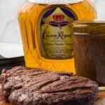 Two grilled steaks rest on a wooden board in front of a glass bottle labeled "Crown Royal" and a jar filled with dark whiskey steak marinade, all set against a light background.
