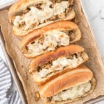 Four toasted hoagie rolls filled with seasoned chicken cheesesteak and topped with melted cheese are arranged on a parchment-lined baking sheet.