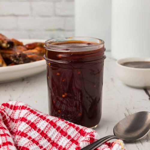 Sweet chili sauce in a glass jar with a plate of grilled ribs and dipping sauce in the background, on a rustic table.