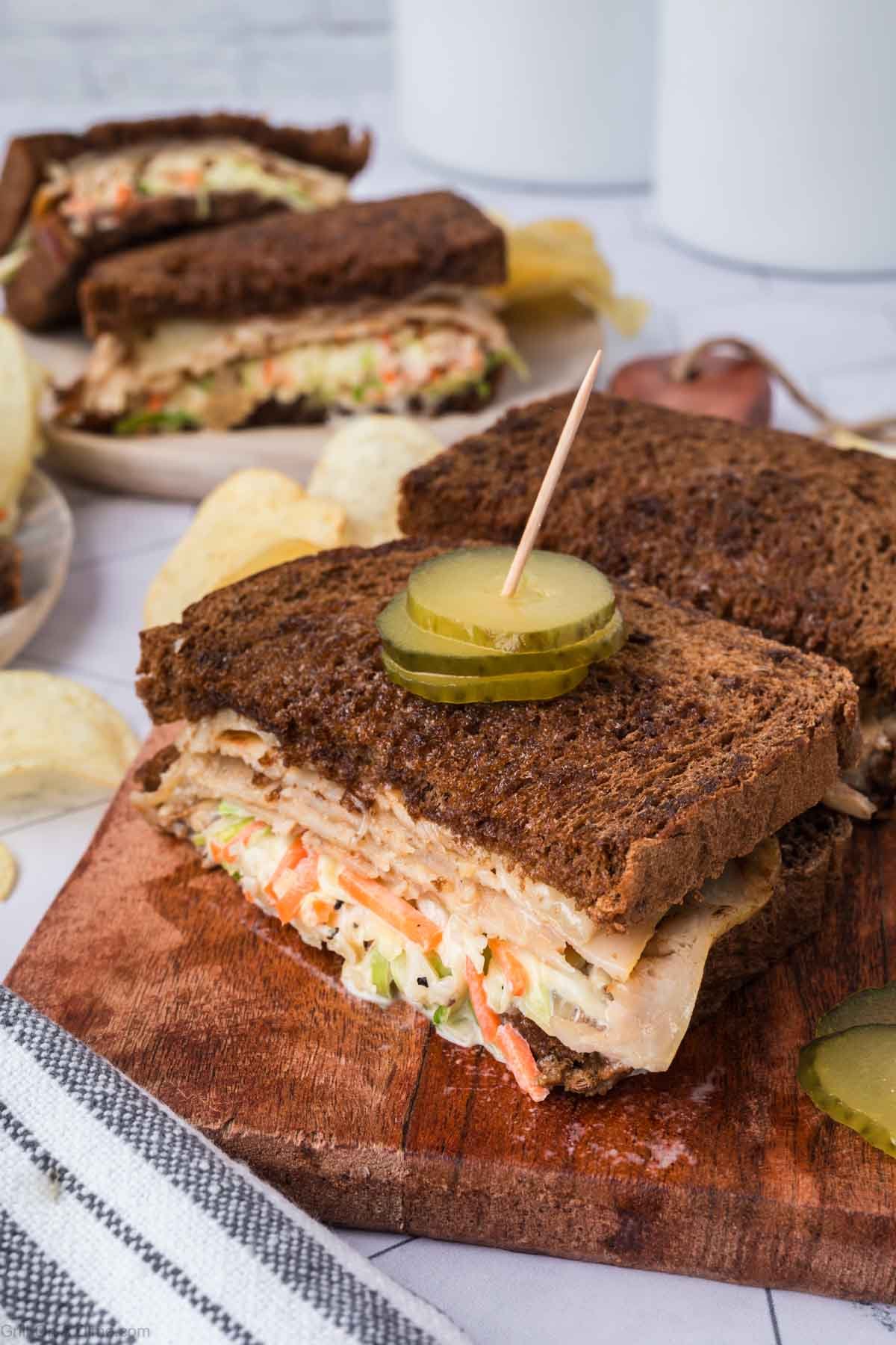 Rye Bread Sandwich cut in half with slices of pickles with a toothpick inserted in them on top with layers of turkey and coleslaw