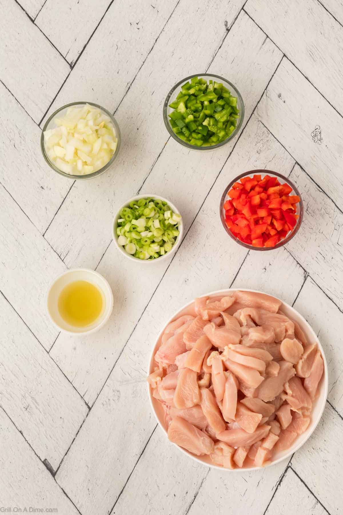 Sliced raw chicken in a bowl with small bowls of diced bell peppers, green onions, white onions and oil