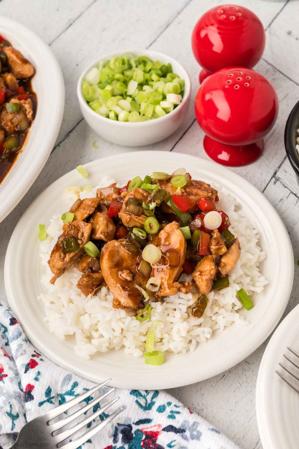 Bourbon Chicken topped on white rice on a plate with a small bowl of green onions on the side