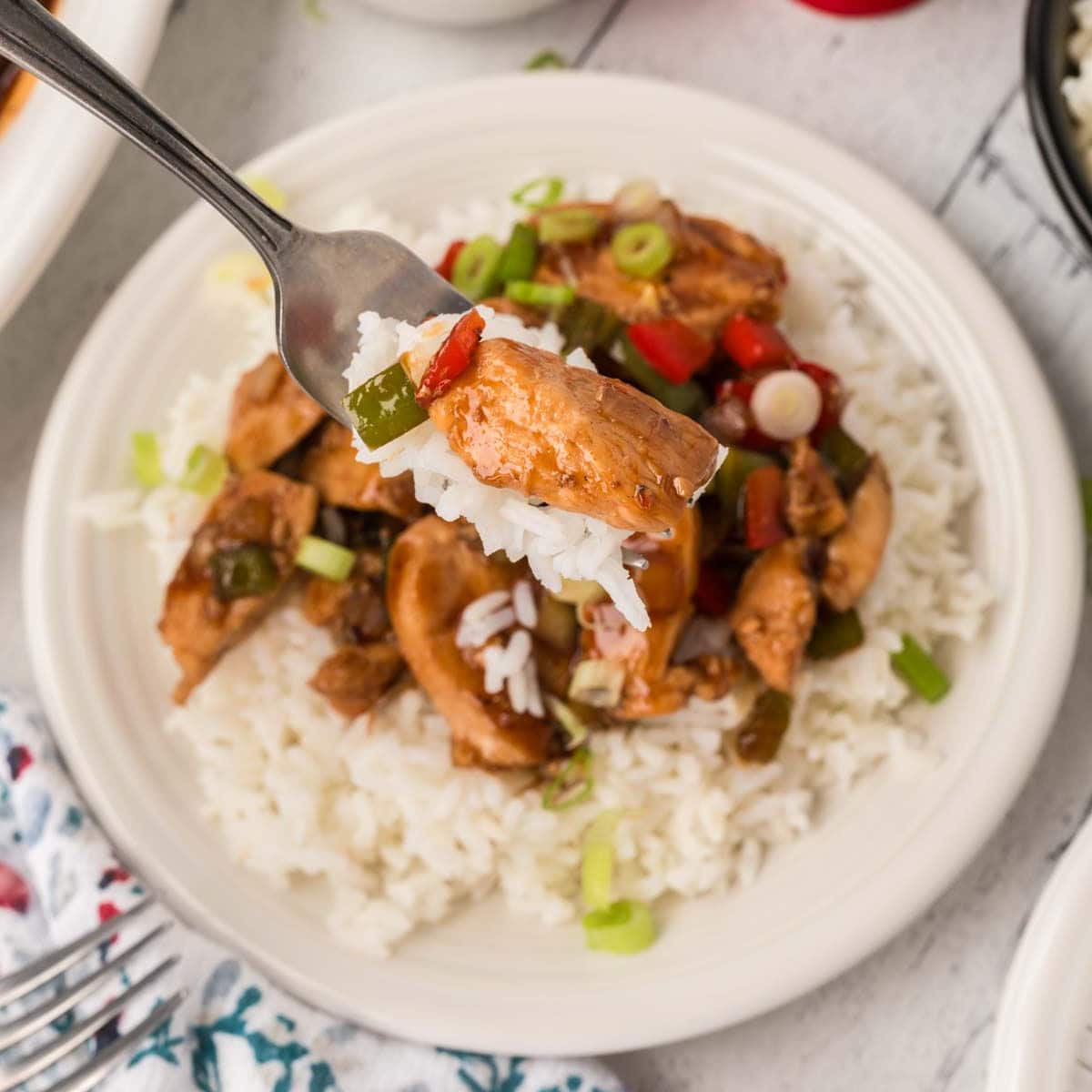 A bite of Bourbon Chicken on a fork with a plate of Bourbon Chicken Mixture over white rice in the background