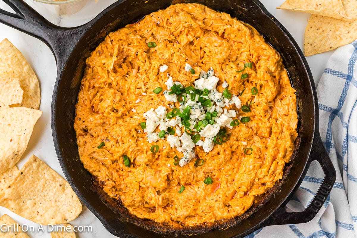 Buffalo Chicken Dip in a cast iron skillet topped with blue cheese crumbles and chopped green onions with a side of tortilla chips