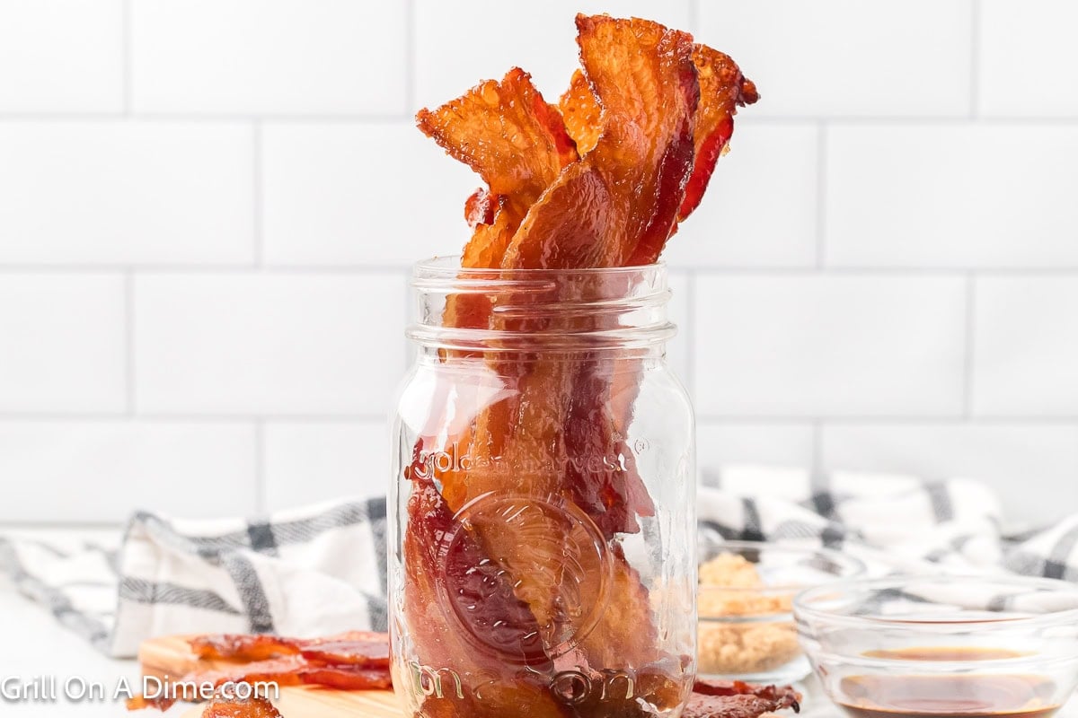 Strips of candied bacon in a jar with small bowls of maple syrup and brown sugar on the side
