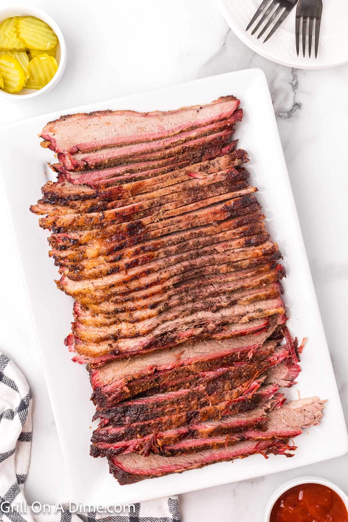 Sliced Brisket on a white platter with a small bowl of slice pickles on the side