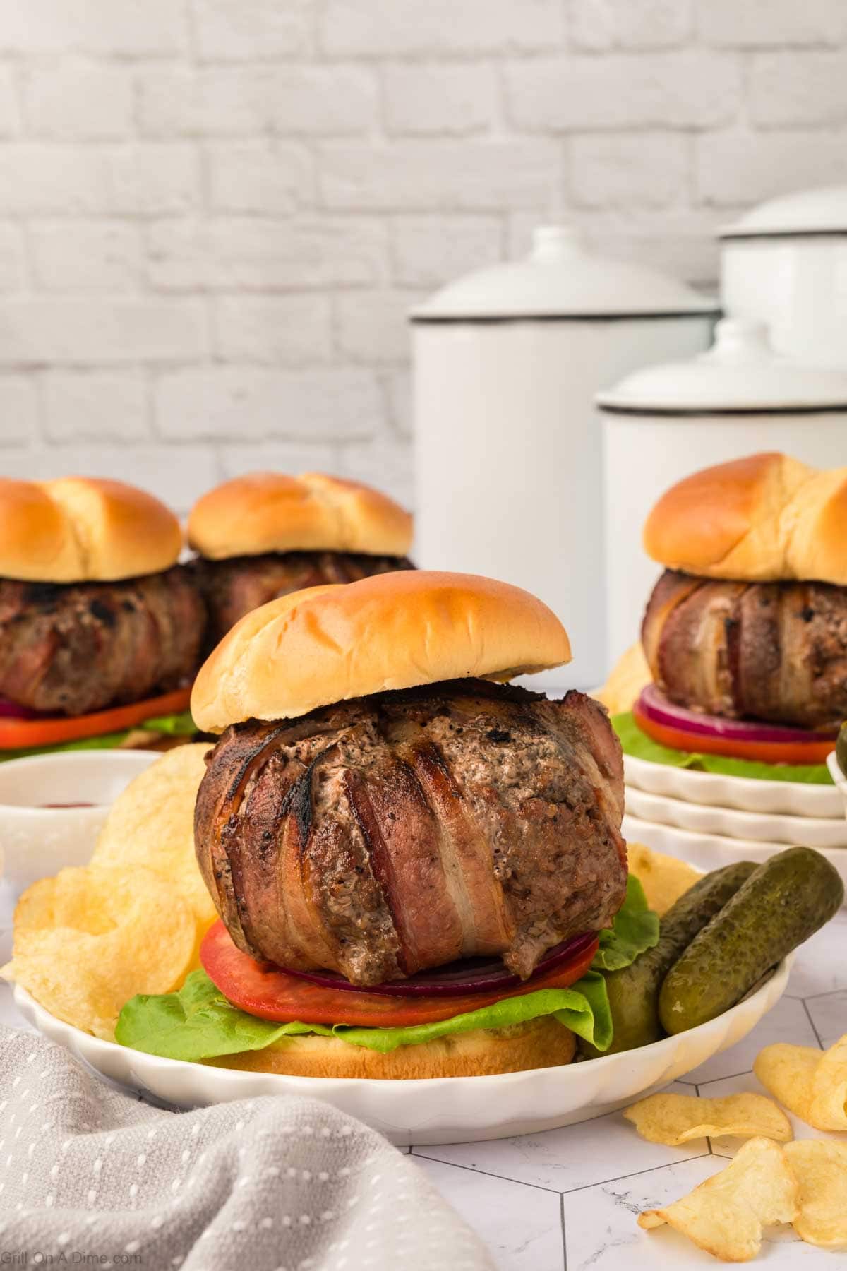 Bacon wrapped burger on a plate with pickles and potato chips