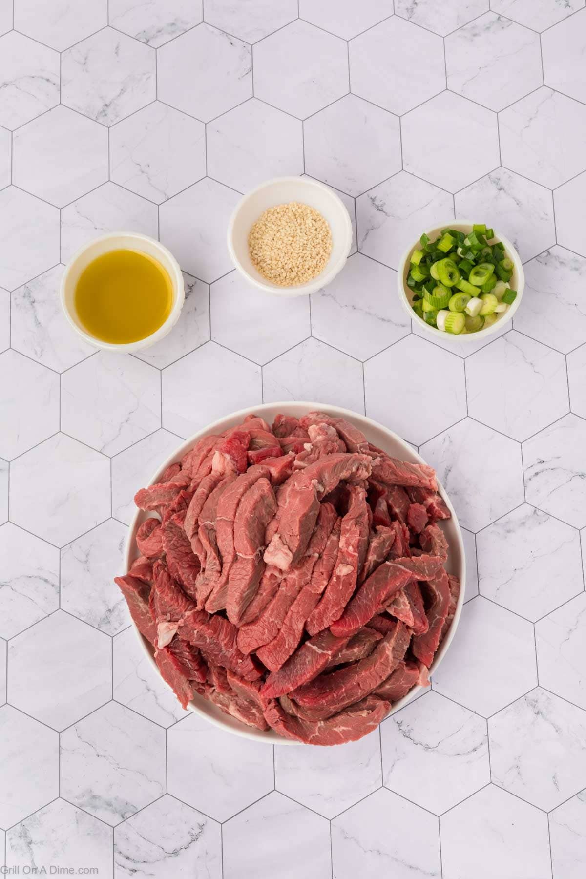 Ingredients - Thinly slices of steak in a bowl with small bowls of oil, sesame seeds and chopped green onions