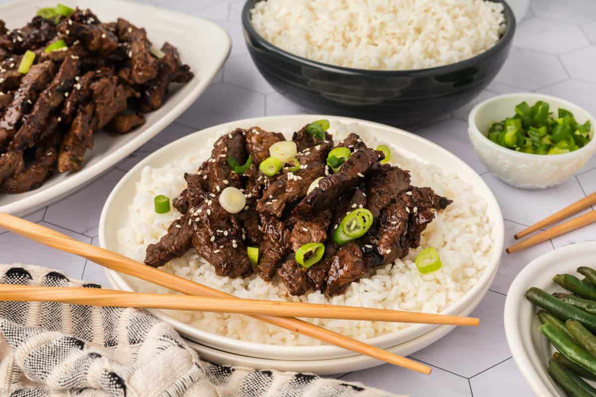 Bowl of white rice and a platter of Korean BBQ Beef on a platter with a serving of the beef over rice topped with green onions