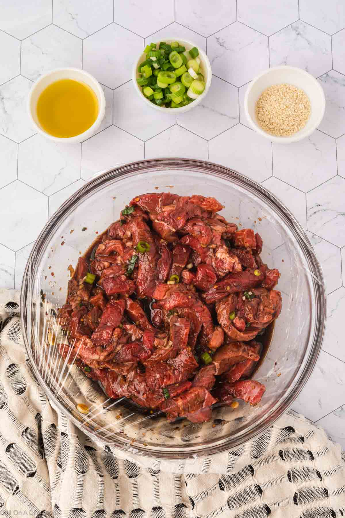 Plastic wrap covering the bowl with the strips of steak and marinade in the bowl with small bowls of oil, green onions and sesame seeds on the side
