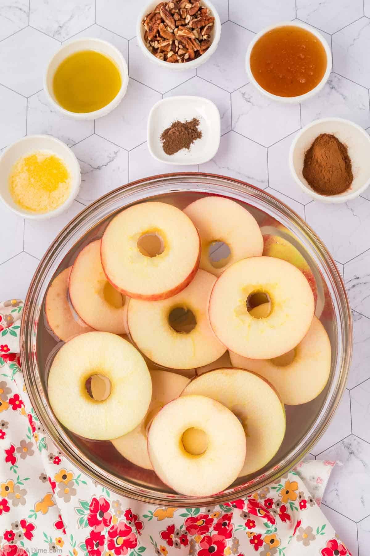 Slice apples in a bowl with small bowls of spices, oil, honey, and chopped pecans