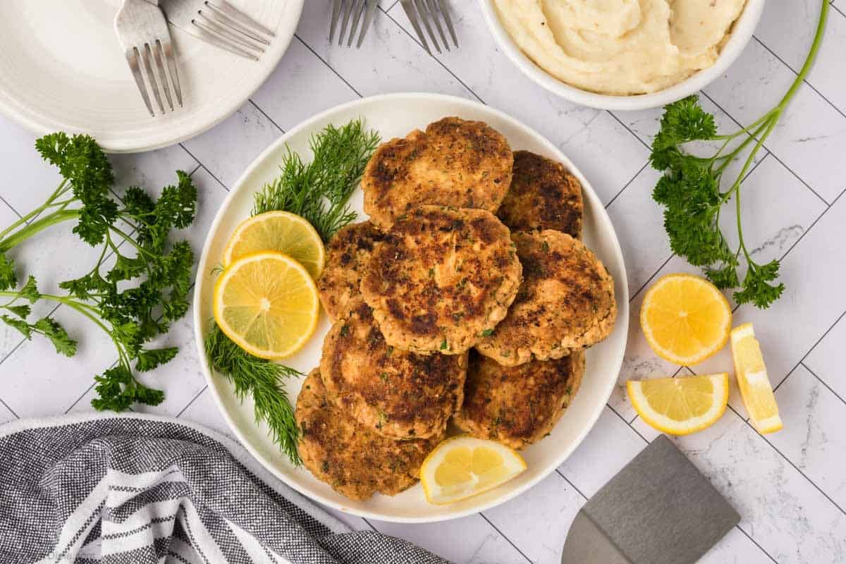 Salmon patties stacked on a white plate with fresh herbs and lemon slices on the side