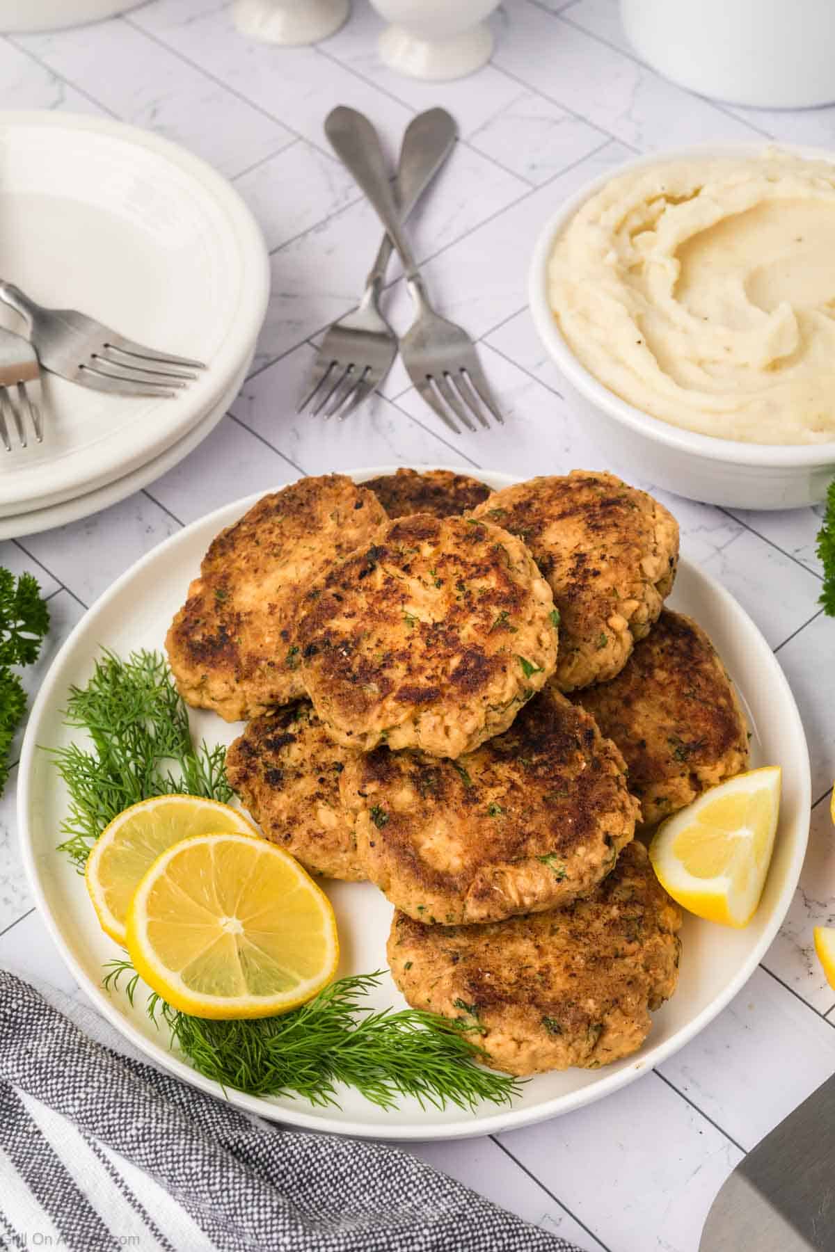 Salmon Patties stacked on a white plate with fresh lemon slices and fresh herbs on the side