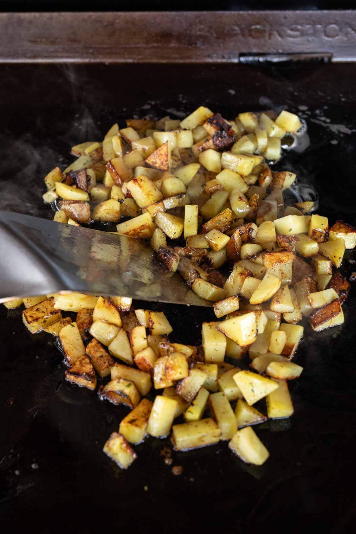 Diced potatoes being cooked and stirred on a flat black griddle with a metal spatula, alongside sizzling Blackstone Bacon Ranch chicken. The potatoes appear golden brown and slightly crispy as steam rises from the hot surface.