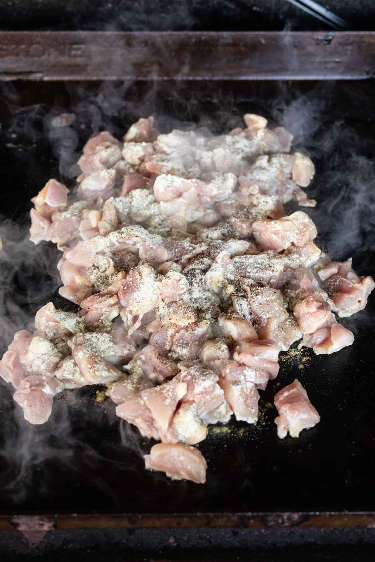 Chopped raw chicken pieces with seasoning are sizzling and steaming on a flat, dark Blackstone griddle, perfect for making flavorful Bacon Ranch Chicken.