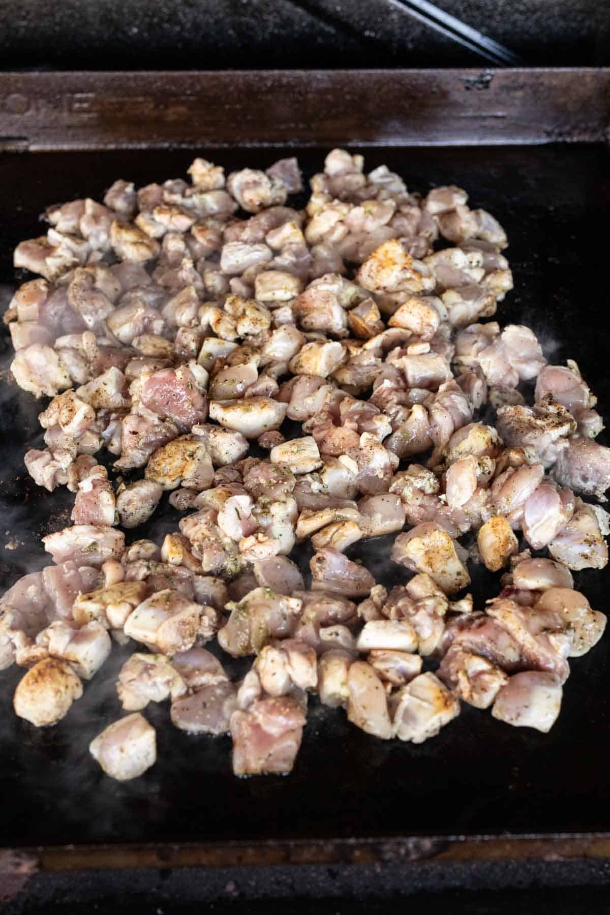 Chopped, seasoned raw Blackstone Chicken pieces cooking on a flat top grill or griddle, with steam rising from the hot surface.