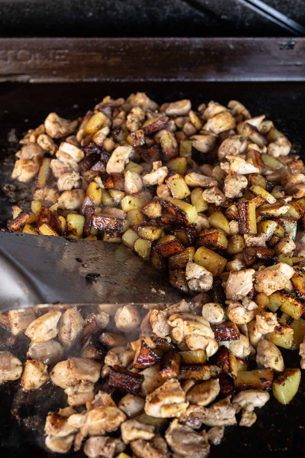 Diced chicken and golden-brown potatoes sizzle on a Blackstone griddle, stirred with a metal spatula. Steam rises as this Chicken Recipe cooks to perfection.