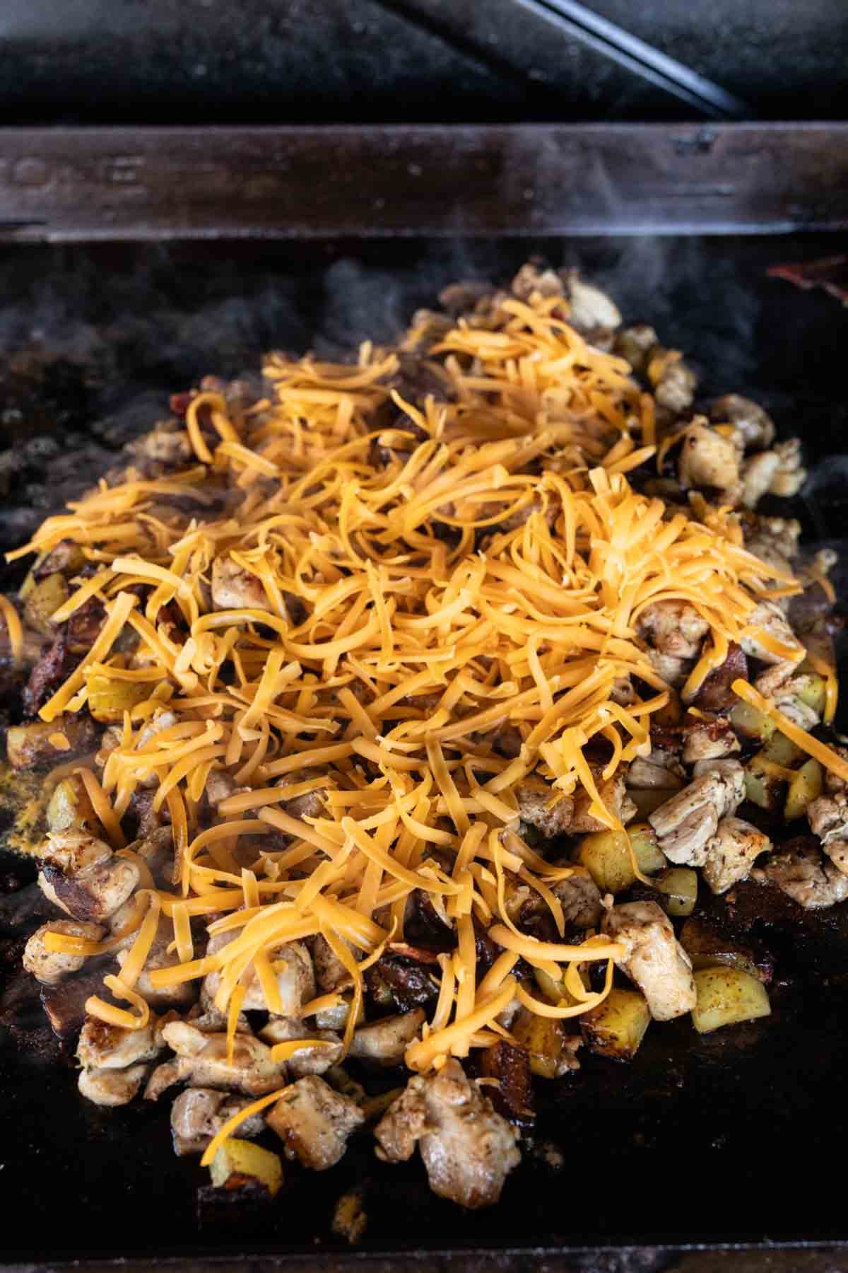 Bacon Ranch Chicken, diced vegetables, and shredded cheddar cheese are sizzling on a Blackstone grill, with steam rising as the cheese melts over the chicken and veggies—a mouthwatering addition to your favorite Blackstone recipes.