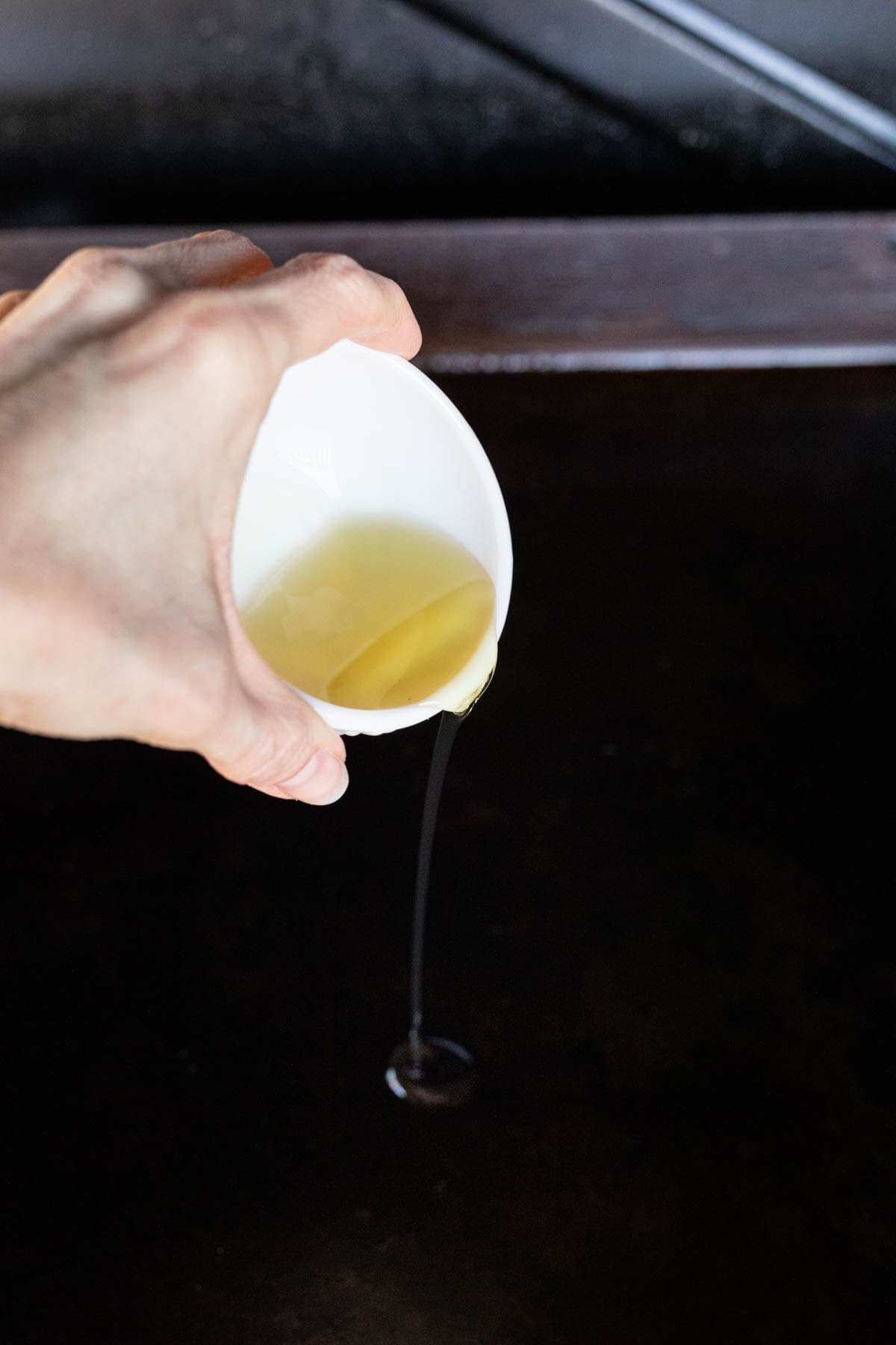 A hand pours oil from a small white cup onto a dark Blackstone cooking surface, preparing it for Bacon Ranch Chicken, with a thin stream of oil falling below.