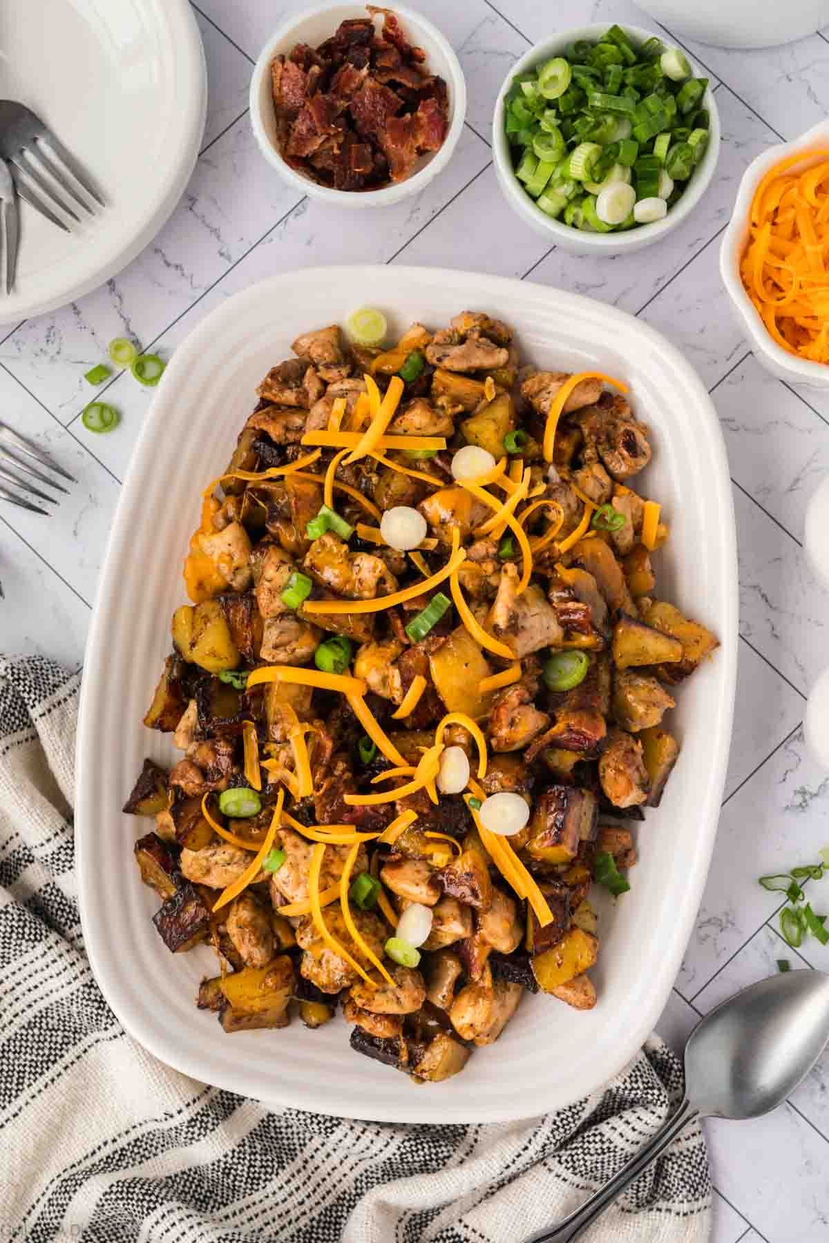 A white platter holds a Bacon Ranch Chicken dish with roasted potatoes, garnished with green onions and shredded cheddar cheese. Surrounding the platter are bowls of bacon bits, green onions, and cheese. A fork and spoon are nearby.