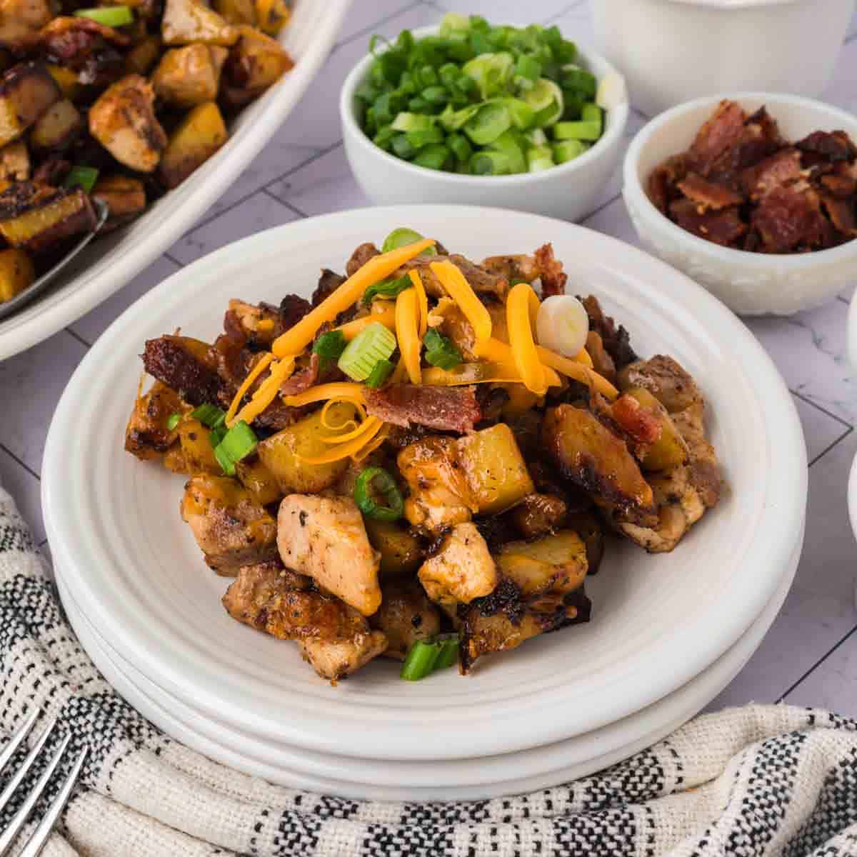 A white plate filled with chunks of seasoned, grilled Blackstone Chicken and potatoes, topped with shredded cheddar cheese, green onions, and crumbled bacon. Small bowls of green onions and bacon bits are in the background.