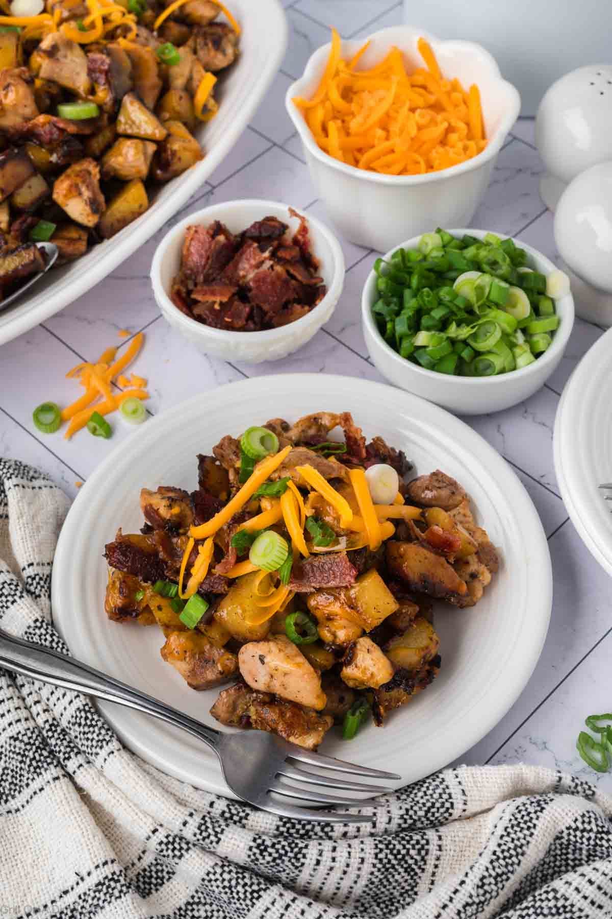 A plate of loaded breakfast hash with potatoes, Blackstone Bacon Ranch Chicken, crispy bacon, shredded cheddar cheese, and green onions, surrounded by small bowls of chopped green onions, shredded cheese, and bacon. A fork and knife rest beside the plate.