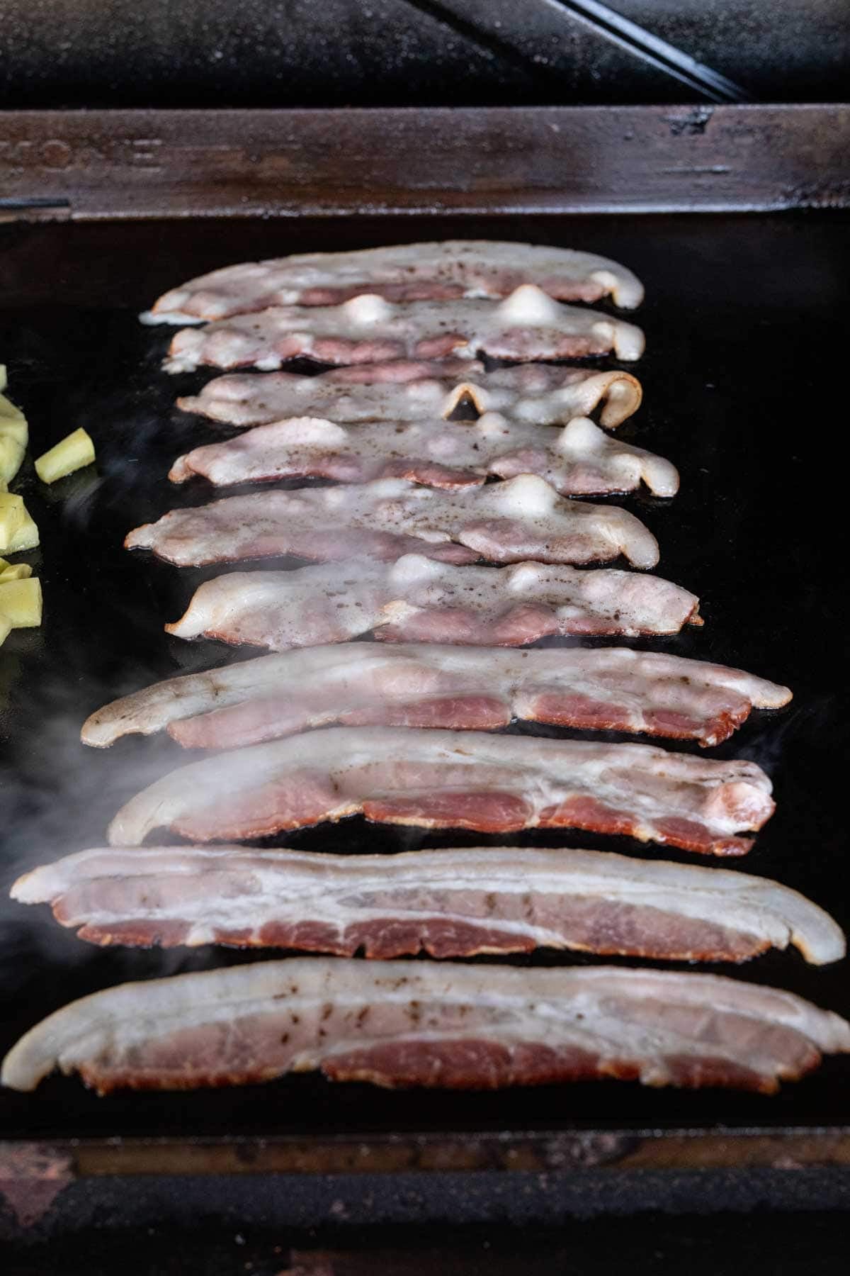 Several strips of bacon sizzle on a Blackstone griddle with steam rising, and a small portion of diced potatoes is visible to the left side—a classic Blackstone recipe setup.