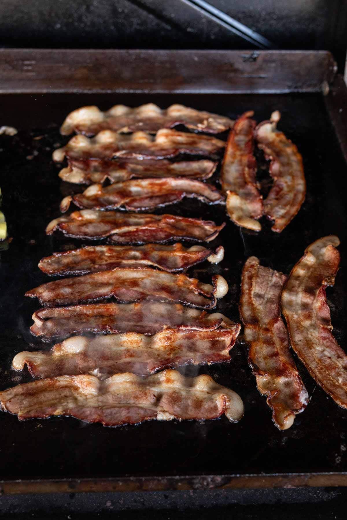 Several strips of bacon are sizzling on a Blackstone griddle, with some pieces crisp and others still soft, surrounded by bubbling grease—a delicious step in preparing a savory Bacon Ranch Chicken recipe.