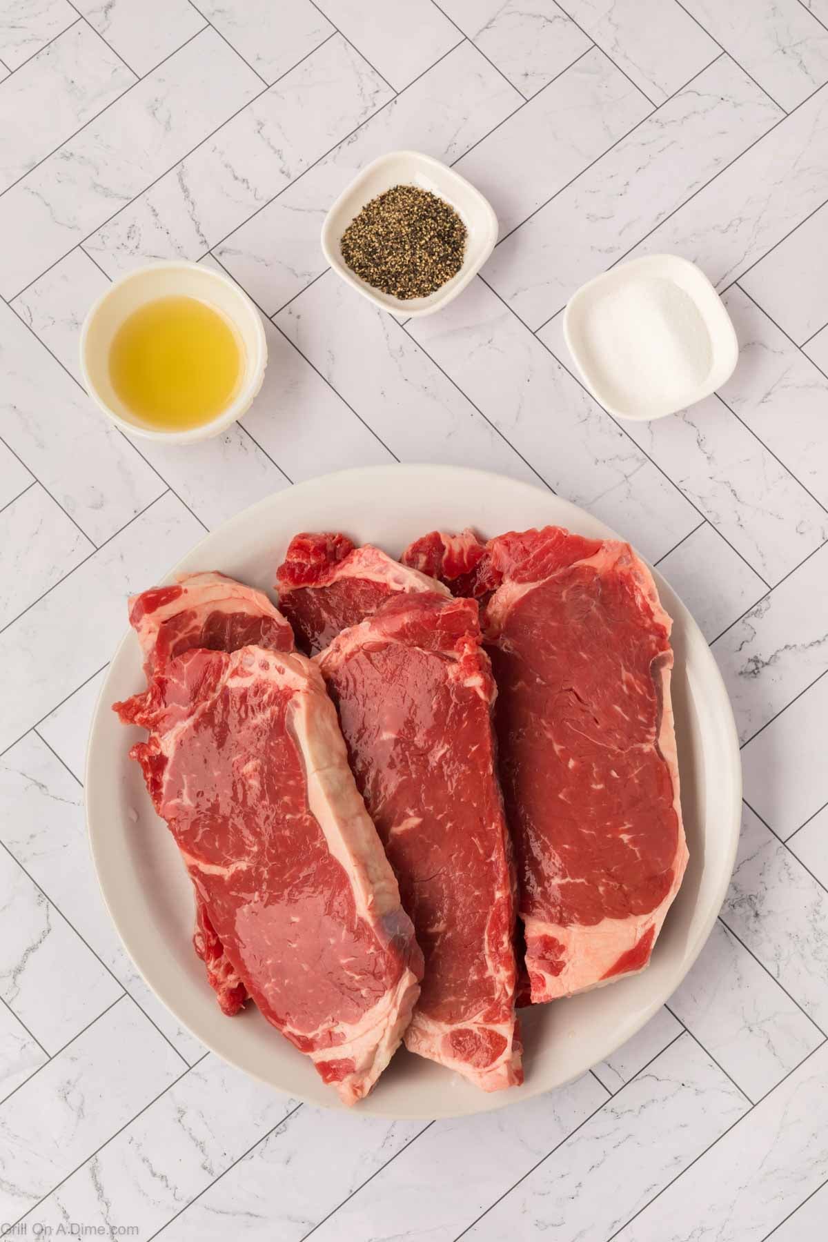 Three raw steaks on a white plate, ready to become a delicious Garlic Butter Steak, are placed next to small bowls of oil, black pepper, and salt on a white tiled surface.