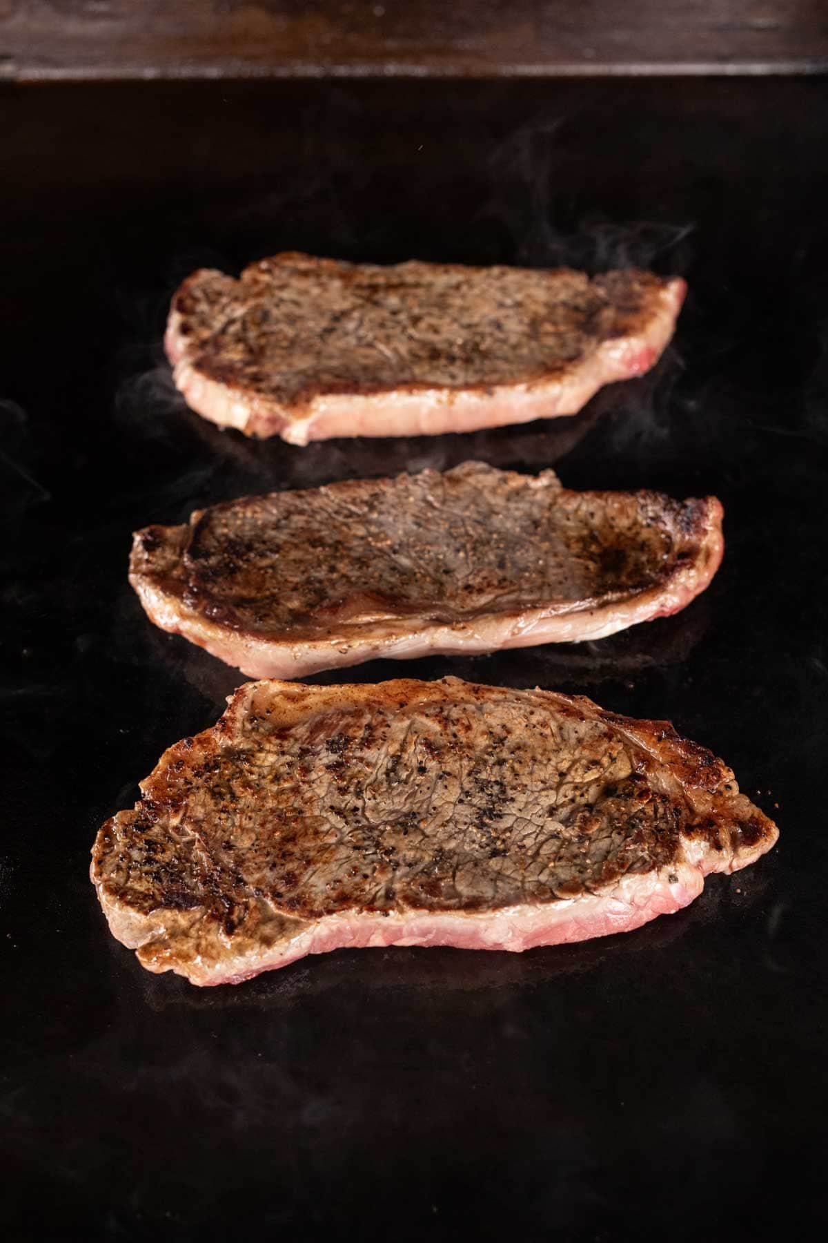 Three seared steaks cooking on a flat, dark griddle surface, with visible browning and steam rising from the meat—a perfect Garlic Butter Steak for fans of Blackstone recipes.