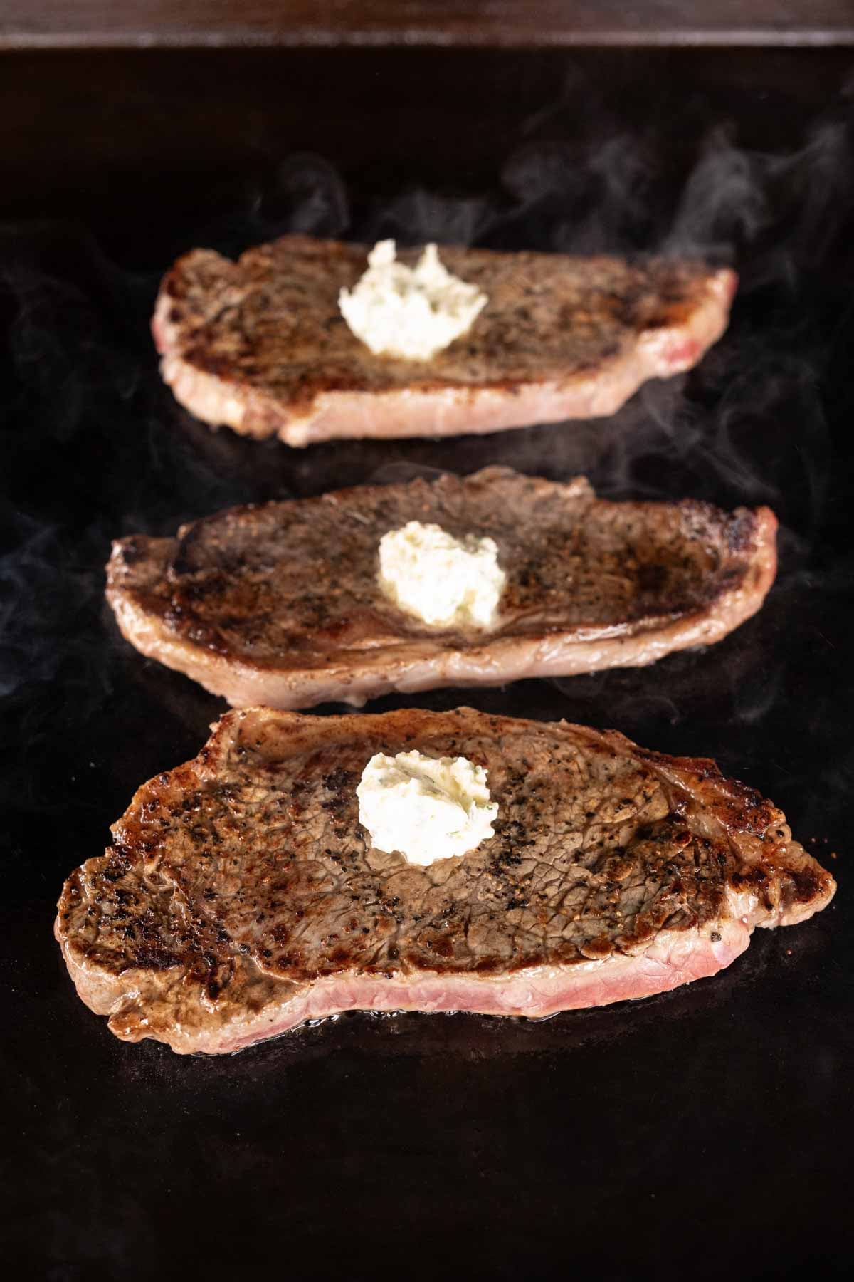 Three cooked Garlic Butter Steaks are lined up on a Blackstone grill, each topped with a dollop of melting compound butter, with steam rising from the hot surface.