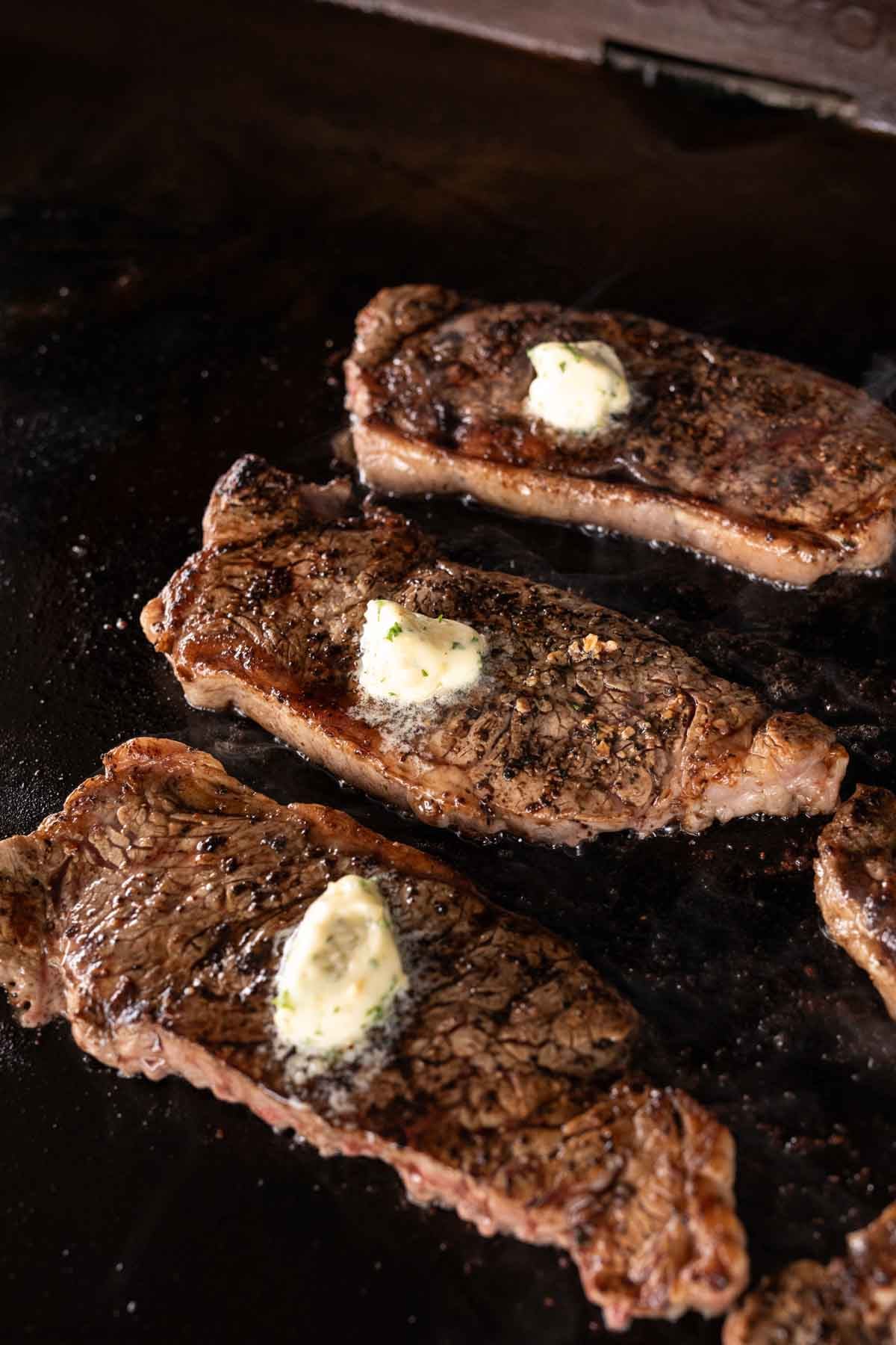 Three cooked Blackstone steaks with seared, seasoned surfaces sit on a dark griddle, each topped with a melting pat of garlic butter.