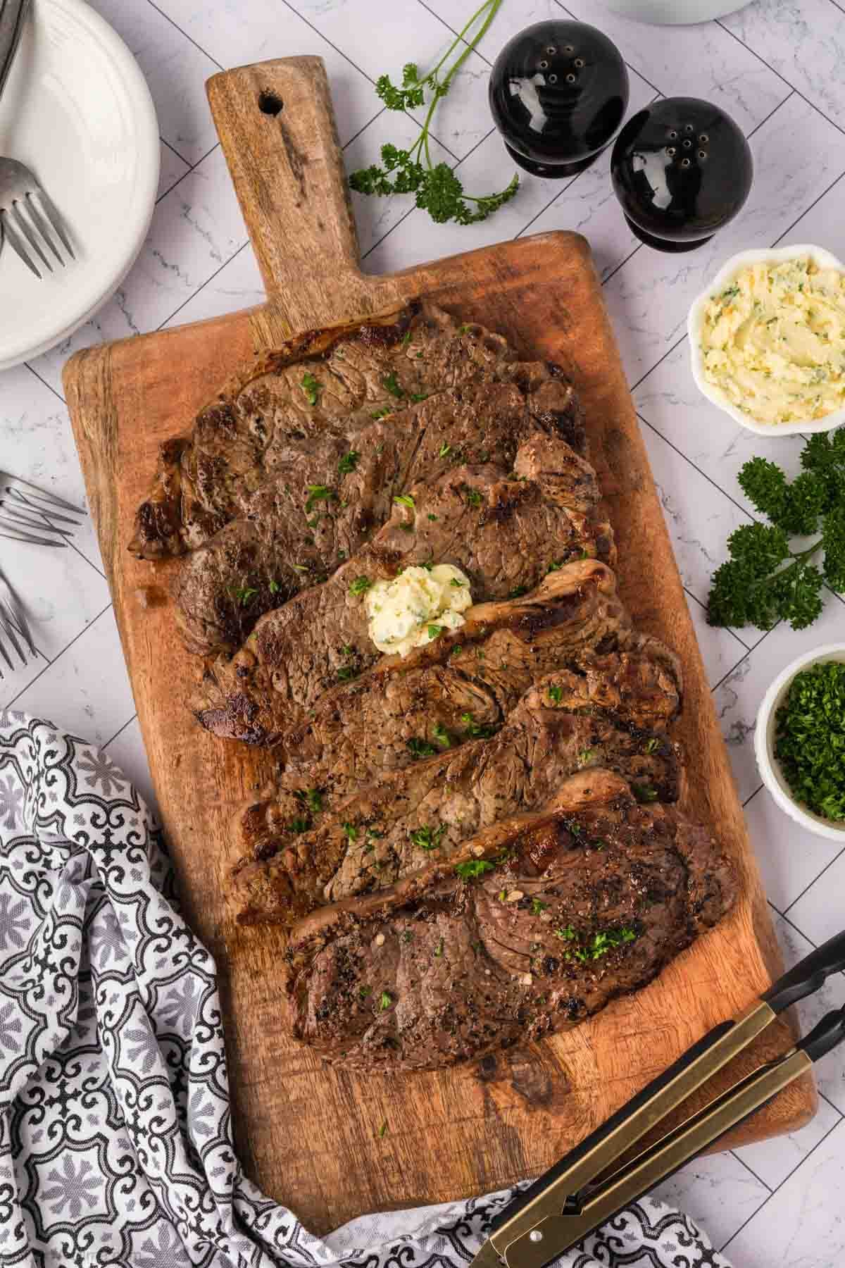 Sliced grilled ribeye steak topped with a dollop of garlic butter on a wooden cutting board, surrounded by fresh parsley, a bowl of butter, forks, and black-and-white napkin—perfect for a classic Garlic Butter Steak night.