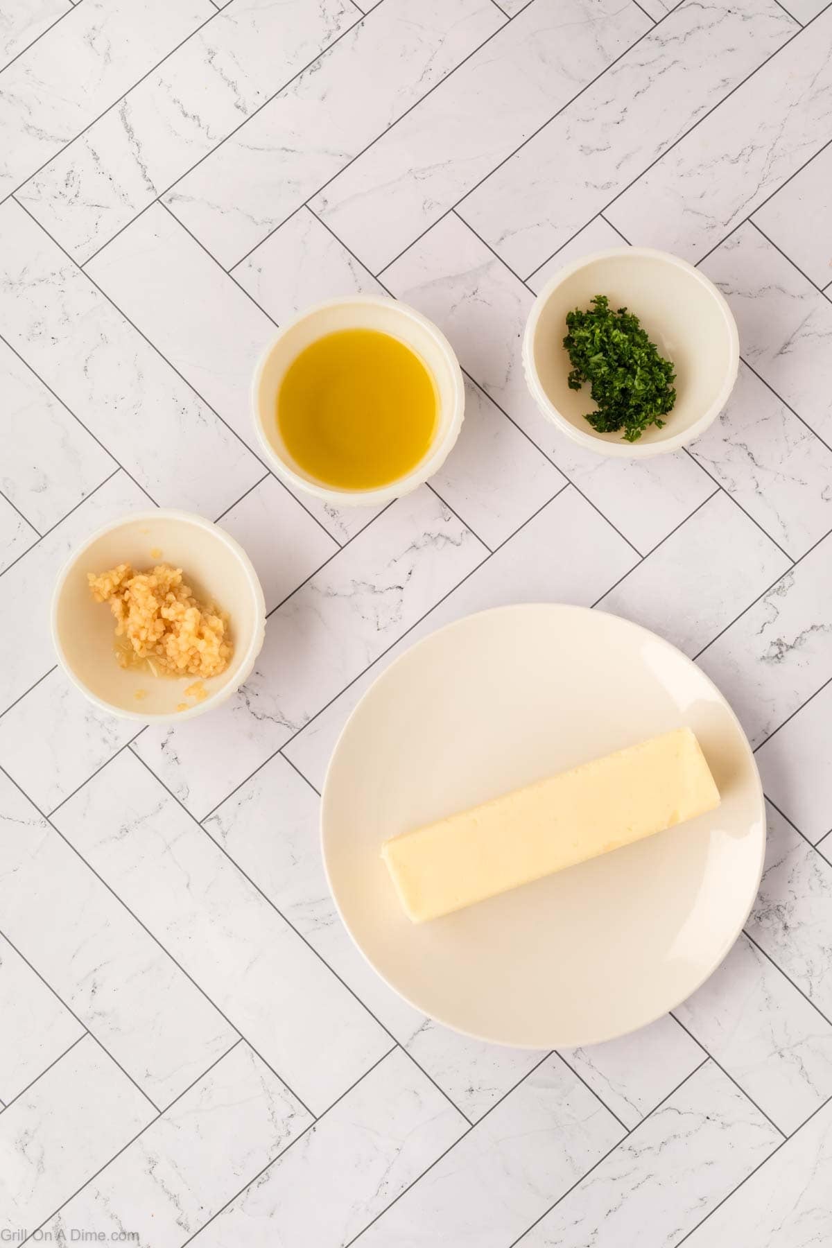 A stick of butter on a white plate, with small bowls of minced garlic, chopped parsley, and olive oil arranged on a tile-patterned surface—perfect for creating flavorful Garlic Butter Steak on your Blackstone.