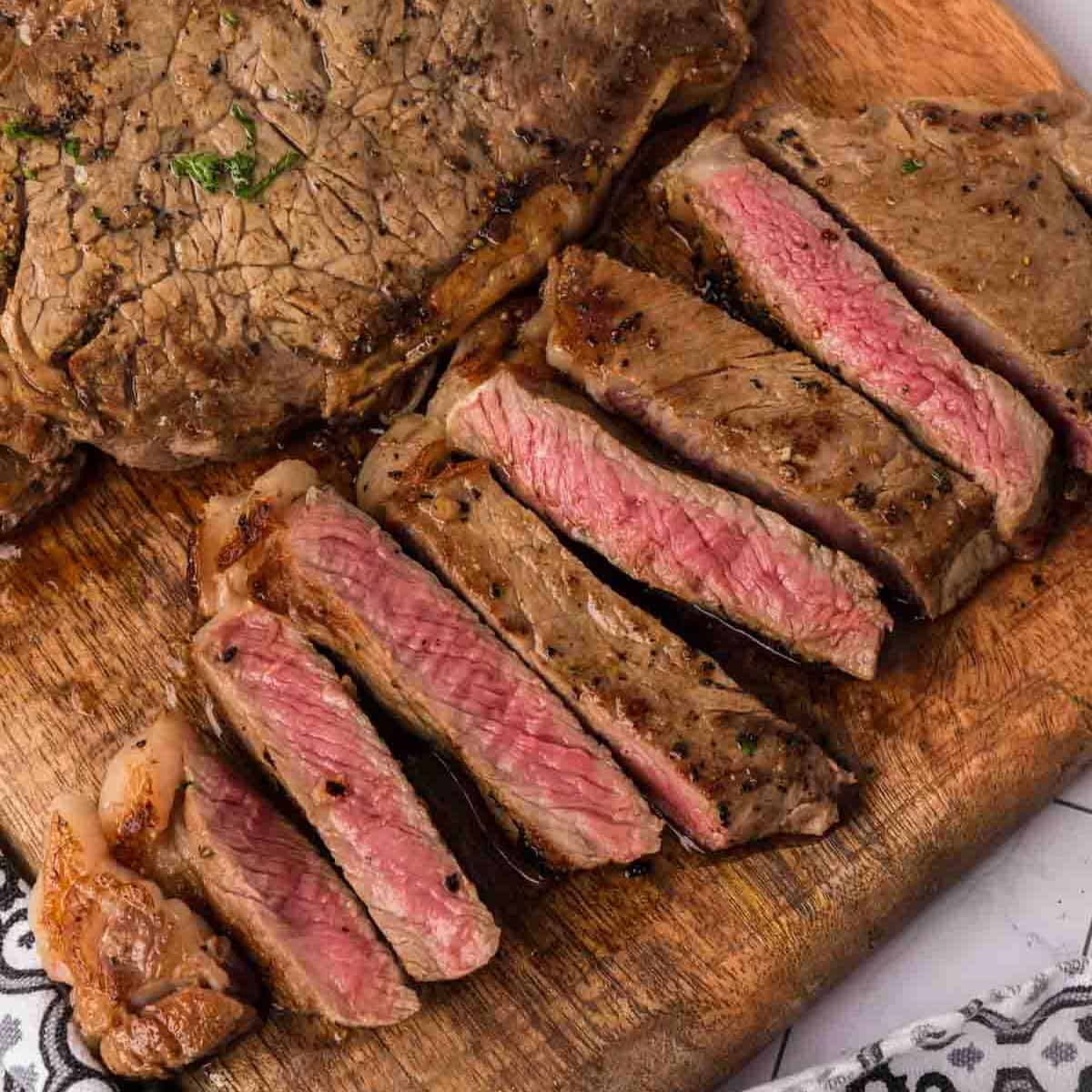 A sliced, medium-rare garlic butter steak is arranged on a wooden cutting board, showing a juicy pink center and seared brown crust—a mouthwatering Blackstone steak recipe with part of the whole steak visible in the background.
