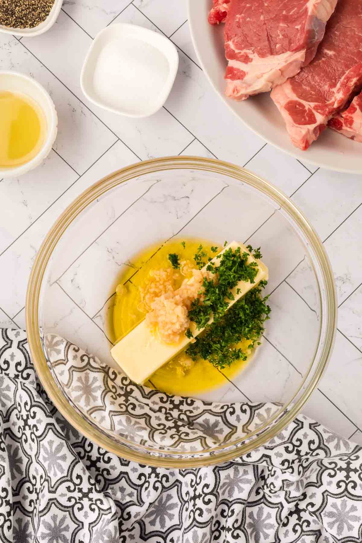 A glass bowl with garlic butter, minced garlic, chopped parsley, and melted butter sits on a white tiled surface. Nearby are salt, oil, black pepper, and a plate with raw steak—perfect prep for Garlic Butter Steak on the Blackstone.