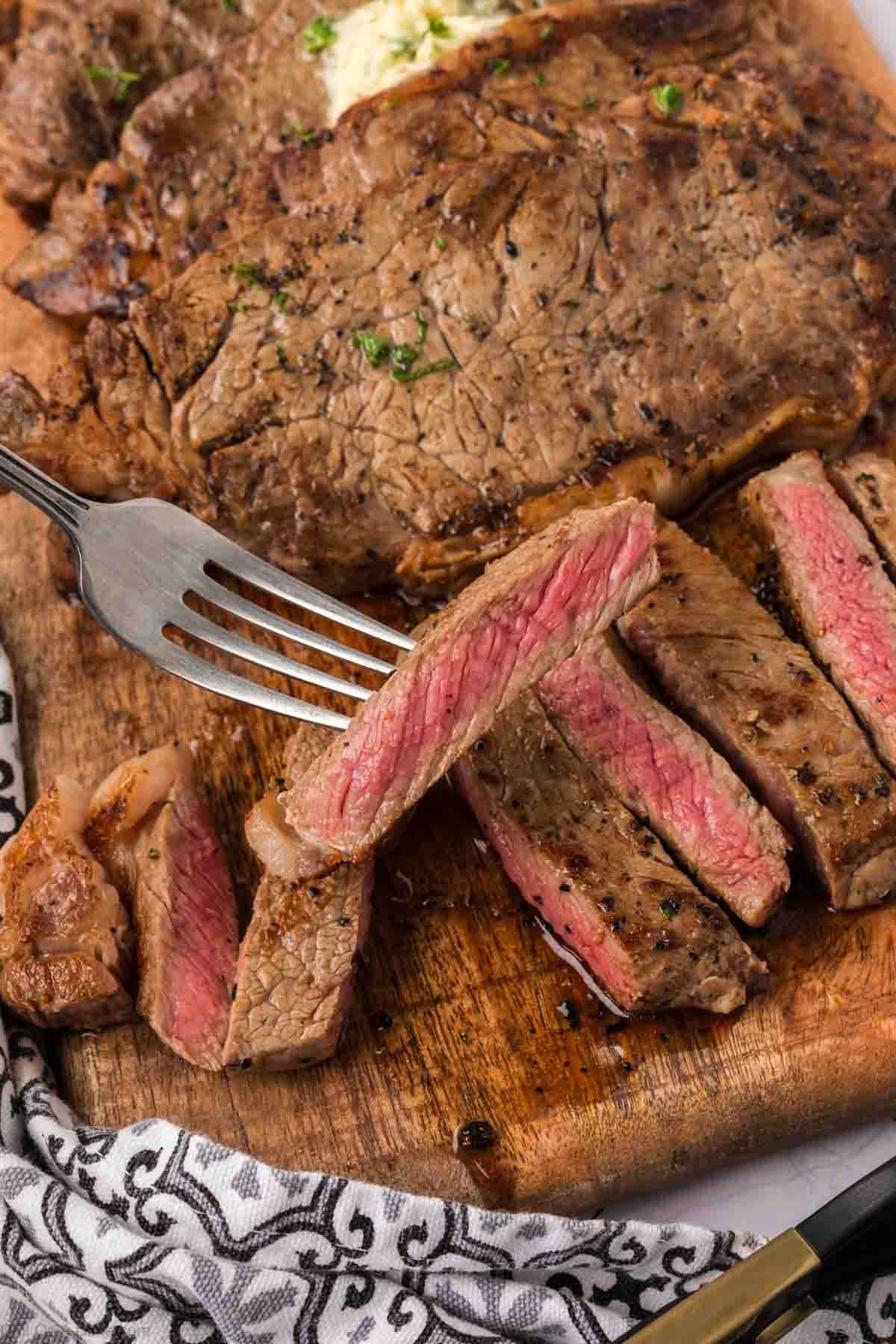 A fork holds a juicy slice of Garlic Butter Steak above a wooden cutting board with more sliced and unsliced pieces, lightly garnished with herbs—a tempting dish perfect for fans of Blackstone recipes. A patterned cloth napkin is nearby.