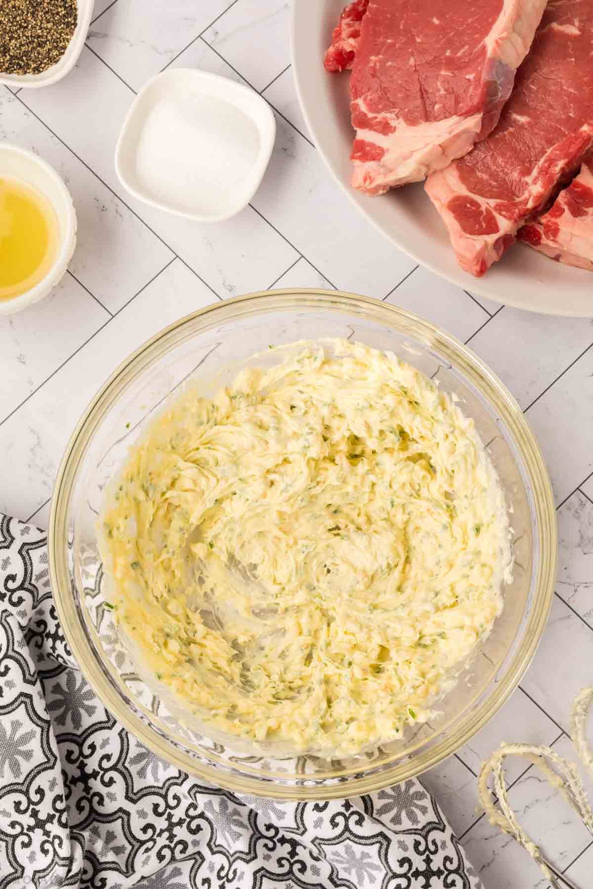 A glass bowl of herbed garlic butter sits on a white counter next to raw Blackstone steak on a plate, a small bowl of melted butter, and salt. A patterned black-and-white cloth is partially visible in the corner.